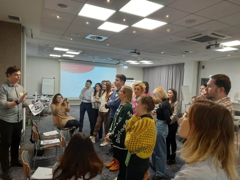 A group of around 20 people stand in an informal semi-circle, listening to a presenter 