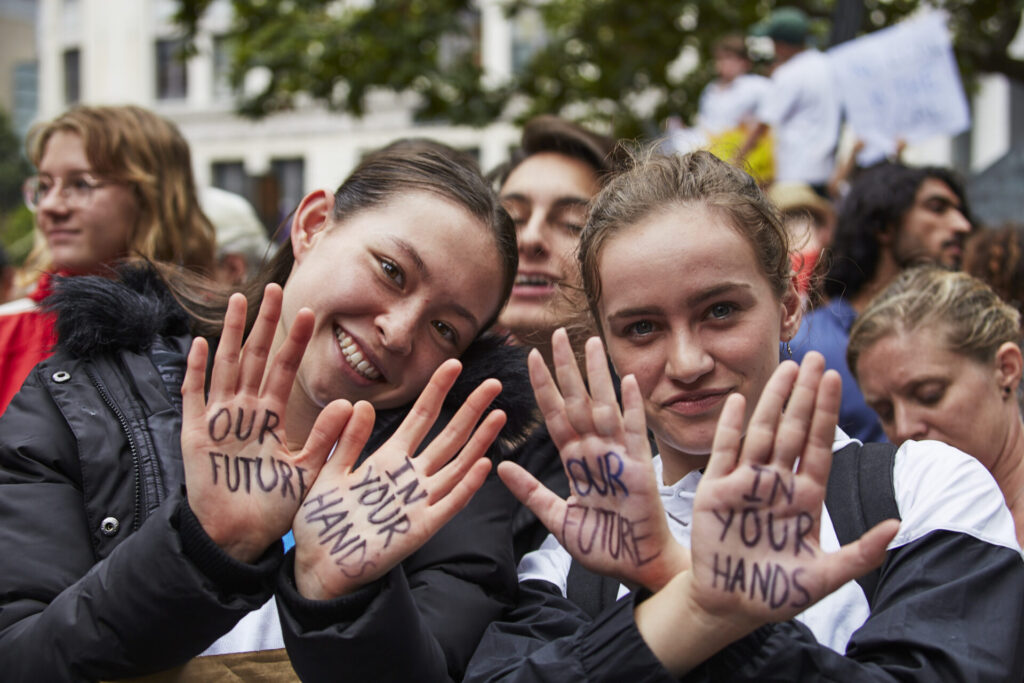 Two girls hold their hands up toward the camera, showing us the message in black texter on their palms "Our future in your hands" behind them is a crowd of people carrying protest signs. 